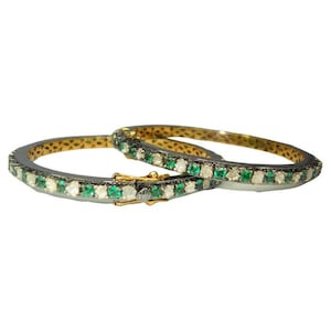 Puede incluir: Dos brazaletes de oro con base de metal negro, adornados con una fila de pequeñas piedras preciosas verdes y blancas.