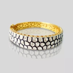 Könnte beinhalten: Ein goldenes Armreif-Armband mit einer Reihe kleiner, weißer, unregelmäßig geformter Diamanten, die in einen schwarzen Metallrahmen gefasst sind.