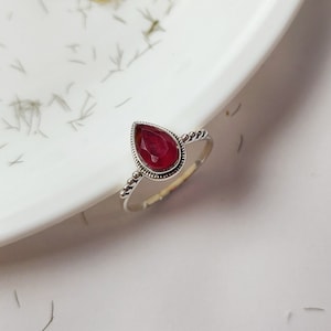 May include: A silver ring featuring a teardrop-shaped red gemstone set in a silver bezel. The ring has a beaded band.