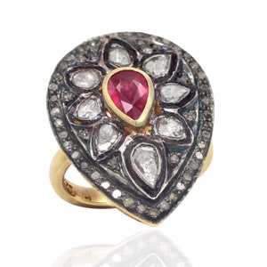 Könnte beinhalten: Ein tropfenförmiger Ring mit einem roten Rubin-Edelstein, der von einer Gruppe kleinerer Diamanten umgeben ist. Der Ring ist in Gold gefasst und hat eine silberfarbene Band.
