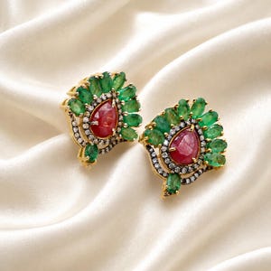 May include: A pair of gold earrings with a central, teardrop-shaped red gemstone, encircled by small white stones and green emeralds. The earrings are displayed on a white, silky surface, highlighting their detailed design and vivid colours.
