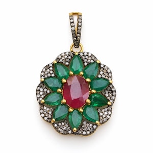 Peut inclure: Un pendentif en forme de fleur avec un rubis rouge ovale central entouré de huit émeraudes vertes en forme de poire. Le bord extérieur est incrusté de petits diamants pavés sur métal sombre, avec une bélière ornée de diamants au sommet.