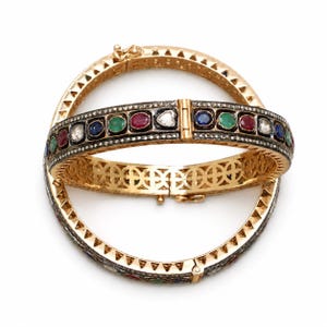May include: Two gold hinged bangles featuring a central row of alternating round-cut red, green, blue, and clear stones set in dark metal. The edges are lined with small pavé stones, and the sides display a geometric triangular cutout pattern.