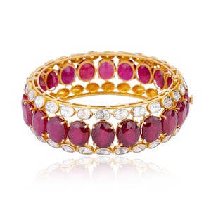 Puede incluir: Un brazalete de oro con tres filas de rubíes rojos y diamantes blancos. El brazalete está sobre un fondo blanco.