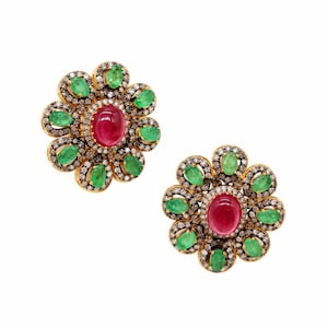 May include: A pair of flower-shaped earrings featuring a central oval-cut red gemstone surrounded by a halo of small green oval-cut stones and a outer border of tiny clear pavé-set diamonds, all set in a gold-toned metal frame.
