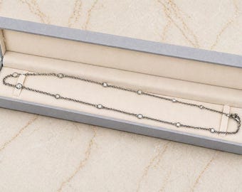 Puede incluir: Un collar plateado con piedras preciosas redondas y transparentes se muestra dentro de una caja de joyería gris con un interior color crema. El collar está dispuesto en línea recta, mostrando su diseño.