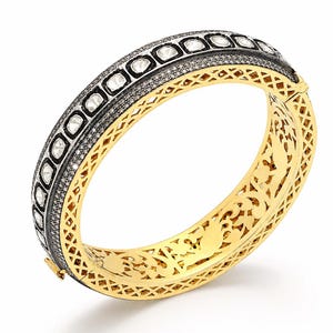 Puede incluir: Un brazalete rígido con bisagra que presenta una banda central de metal oscuro incrustada con pequeñas piedras pavé y una fila de piedras más grandes, claras y de talla rosa. Los lados y el interior son de oro amarillo pulido con un intrincado patrón de celosía.