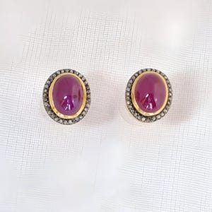 May include: A pair of oval-shaped earrings with a cabochon ruby gemstone set in a gold bezel. The bezel is surrounded by a halo of small diamonds.
