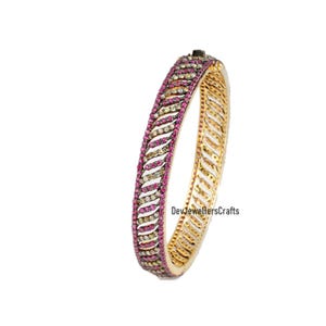 May include: A gold bangle bracelet featuring a design of alternating pink and white gemstones. The bracelet is set with small, round stones and has a polished finish.  "DevJewellersCrafts" is printed on the bracelet.