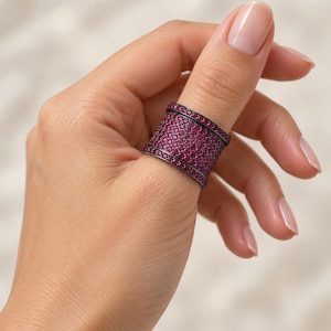 Puede incluir: Un anillo de banda ancha con acabado en metal oscuro y un engaste pavé denso de numerosas piedras circulares pequeñas de color rosa vibrante que cubren toda la superficie, enmarcado por dos filas delgadas de piedras rosas a juego en los bordes.