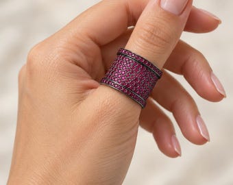 Puede incluir: Un anillo de banda ancha con acabado en metal oscuro y un engaste pavé denso de numerosas piedras circulares pequeñas de color rosa vibrante que cubren toda la superficie, enmarcado por dos filas delgadas de piedras rosas a juego en los bordes.