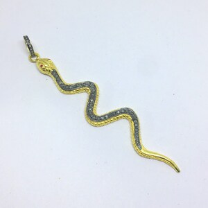 Puede incluir: Un colgante de serpiente de oro con diamantes negros. La serpiente está enroscada en forma de 'S' con la cabeza levantada. El colgante mide aproximadamente 5 cm de largo.