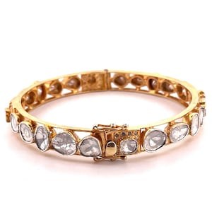 Puede incluir: Brazalete rígido dorado con piedras preciosas claras de forma irregular. El brazalete tiene un cierre seguro y está engastado con piedras de acento claras más pequeñas. El diseño es ornamentado y elegante, adecuado para ocasiones especiales.