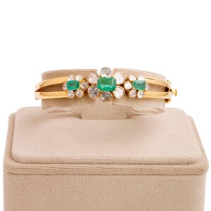 May include: A gold bangle bracelet with a floral design featuring emerald green and clear gemstones. The bracelet is displayed on a beige velvet stand.