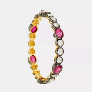Puede incluir: Un brazalete dorado con piedras preciosas de rubí ovaladas y piedras preciosas blancas ovaladas alternas. El brazalete tiene un engaste de metal oscuro alrededor de cada piedra y una banda dorada.