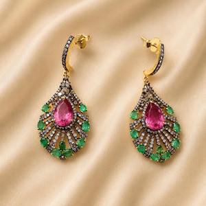 May include: A pair of gold-coloured drop earrings. Each earring features a teardrop design with a large pink gemstone at the centre, surrounded by smaller green gemstones and white accents. The earrings have a post-back closure.