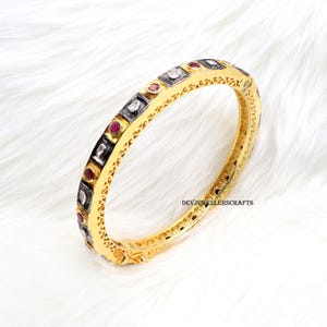 May include: Gold bangle bracelet with a silver and red stone design. The bracelet is decorated with a repeating pattern of small squares and diamonds.
