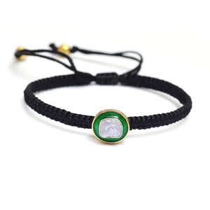 May include: A black braided bracelet with a round, green-enameled pendant featuring a white gemstone.