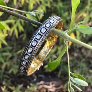 Puede incluir: Un brazalete de oro con una fila de diamantes de forma cuadrada engastados en una banda de metal negro. El brazalete está en una rama verde.