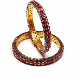 May include: Two gold bangles with a black oxidized finish and a row of red ruby stones and smaller clear stones.