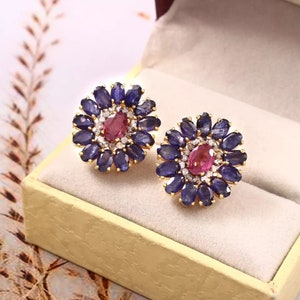 Może przedstawiać: Para złotych kolczyków z kwiatowym wzorem. Kolczyki są ozdobione niebieskimi szafirami i różowym rubinem w środku każdego kwiatu.