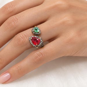Pode incluir: Um anel de estilo aberto com duas pedras preciosas em forma de pera, uma vermelho vibrante e outra verde brilhante, cada uma rodeada por um halo de pequenas pedras claras cravadas numa banda de metal escuro com design ajustável.