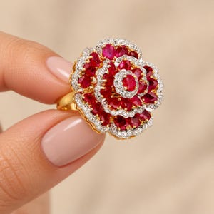 May include: A gold ring featuring a large, multi-layered floral design. The petals are encrusted with numerous oval-cut red rubies and bordered by small, clear white stones, creating a vibrant, layered bloom effect on a polished gold band.