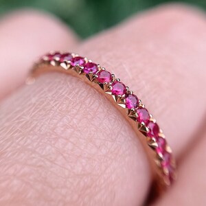 Op de afbeelding: Een roségouden ring met een band van kleine, helderrode edelstenen. Het ontwerp van de ring heeft gelijkmatig verdeelde, rond geslepen stenen, wat een sprankelende, elegante uitstraling creëert. De close-up toont de details van de zetting en de edelstenen.