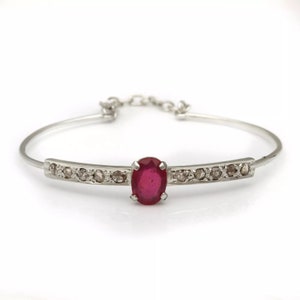 May include: A silver bangle bracelet with a red oval gemstone surrounded by smaller white gemstones.