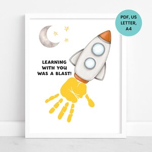 May include: A framed print featuring a white rocket ship with orange accents, a yellow handprint, and the text "LEARNING WITH YOU WAS A BLAST!". The artwork includes a crescent moon and stars.