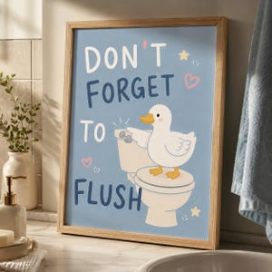 May include: A framed wall art print with a light blue background featuring a white duck standing on a toilet. The text reads DON'T FORGET TO FLUSH in dark blue letters. The illustration includes small pink hearts, yellow stars, and a white toilet.