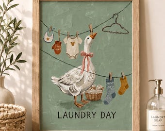 May include: A framed wall art print featuring a white goose with a pink ribbon around its neck, standing in front of a clothesline with baby clothes, socks, and a basket. The text LAUNDRY DAY is printed in black at the bottom on a sage green background.