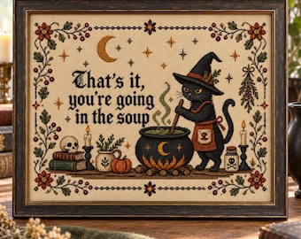 May include: A framed cross-stitch embroidery featuring a black cat in a witch hat and apron stirring a cauldron. The text reads, "That's it, you're going in the soup." The design includes a skull, books, candles, a pumpkin, and floral borders.