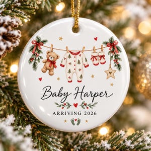 May include: A round white ceramic Christmas ornament featuring a clothesline illustration with a teddy bear, a baby onesie with tree patterns, and booties. The text reads Baby Harper and ARRIVING 2026, accented with red berries, green pine branches, and small gold stars.