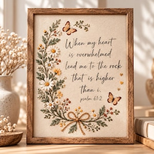 May include: A rectangular wooden frame containing a cream-colored fabric panel with embroidered white daisies, green leaves, and two orange butterflies. The text reads: When my heart is overwhelmed lead me to the rock that is higher than i. psalm 61:2.