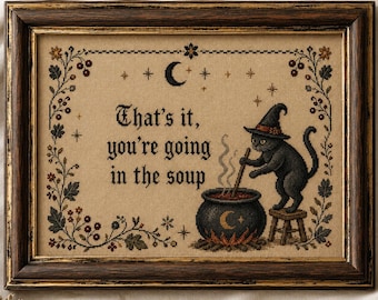 May include: A framed cross-stitch embroidery on tan fabric featuring a black cat in a witch hat stirring a steaming cauldron. The text reads, "That's it, you're going in the soup." The design is bordered by floral vines, stars, and a crescent moon.
