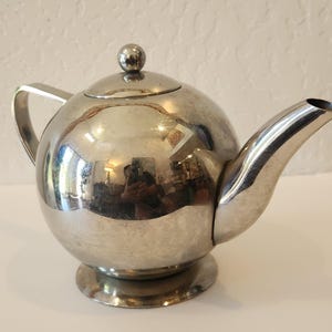 May include: A spherical silver-toned metal teapot with a polished reflective surface. The teapot features a curved handle, a tapered spout, a circular base, and a removable lid topped with a small spherical knob. The metal body is smooth and shiny.
