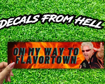 Könnte beinhalten: Ein roter und orangefarbener Aufkleber mit Flammen und dem Text "On My Way To Flavortown" in blau und weiß. Im Hintergrund ist ein Mann mit blonden Haaren und Sonnenbrille zu sehen.