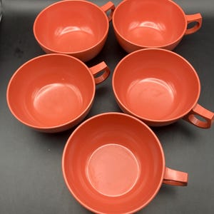 May include: Five coral-colored soup bowls with handles are arranged on a dark surface. The bowls are round with a simple, classic design. The handles are rectangular and attached to the side of each bowl.