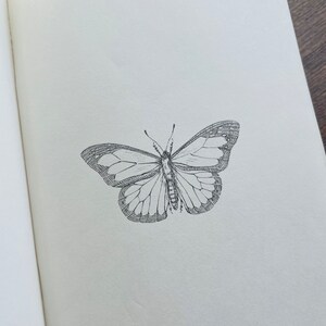 以下が含まれることがあります： 開いた本のページの中央に配置された、詳細な白黒の蝶のイラスト。 蝶の羽は複雑な模様と質感を示し、繊細な静脈とエッジに焦点を当てています。 本のページは柔らかいオフホワイトです。