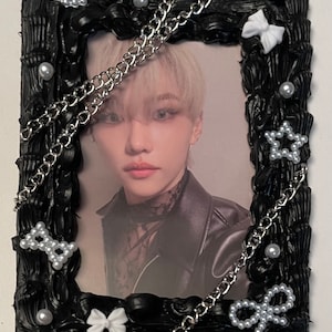 May include: Black picture frame with a white border and silver chains. The frame is decorated with white bows and pearl-like beads. A photo of a person with blonde hair is inside the frame.