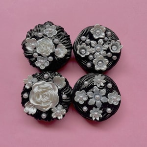 May include: Four black round ornaments with white and silver floral designs. The ornaments have a textured surface and are decorated with pearl-like accents.