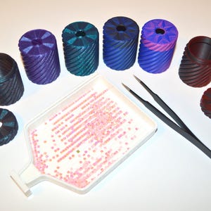 May include: An assortment of colorful, cylindrical containers and circular objects with a textured, ridged design. A white tray holds pink rhinestones, and a pair of black tweezers is also present. The containers are in shades of black, purple, teal, blue, and red.
