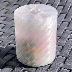 May include: A clear, cylindrical container with a ridged, iridescent design. The container is filled with colorful, small objects. The top of the container has a circular opening. The container is sitting on a dark gray, textured surface.