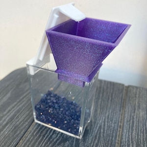 May include: A clear, rectangular container filled with dark blue stones, topped with a purple glittery funnel and a white lid. The funnel is angled and has a square opening. The lid is white and attached to the container.