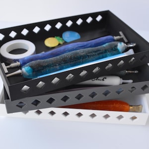 May include: A stack of three rectangular trays in black, gray, and white. The trays have diamond-shaped cutouts. The top tray holds a roll of tape, blue and black glass tubes, and small decorative items. The middle tray contains a white tube with the word "Ink".