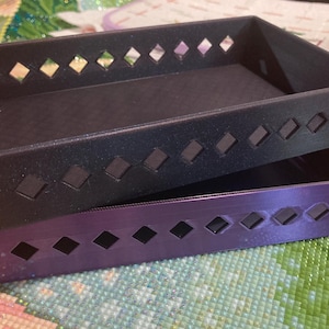May include: Two rectangular storage trays, one stacked on top of the other. The top tray is black, and the bottom tray is purple. Both trays have diamond-shaped cutouts along their sides. The trays are on a colorful, patterned surface.