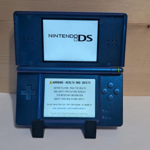 May include: A metallic blue Nintendo DS handheld game console with dual screens. The top screen displays the Nintendo DS logo, while the bottom touch screen shows a health and safety warning message with text instructions to touch the screen.