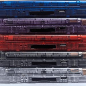 May include: A stack of six transparent plastic cases in various colors: blue, purple, red, gray, and clear. Each case has a rectangular shape with a slot and a USB-C port. The cases are stacked vertically.