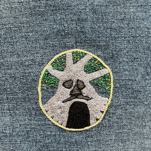 May include: Embroidered patch of a tree with a face, the tree is gray and the face is brown, the tree is surrounded by a yellow circle with green and gold leaves.